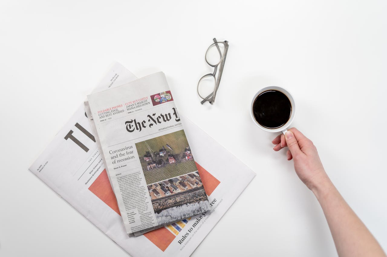 Top view of a coffee cup, newspapers, and eyeglasses on a clean white desk, ideal for a morning routine theme.