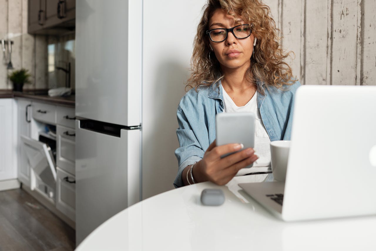 Woman using smartphone and laptop while working remotely in a home kitchen setting.